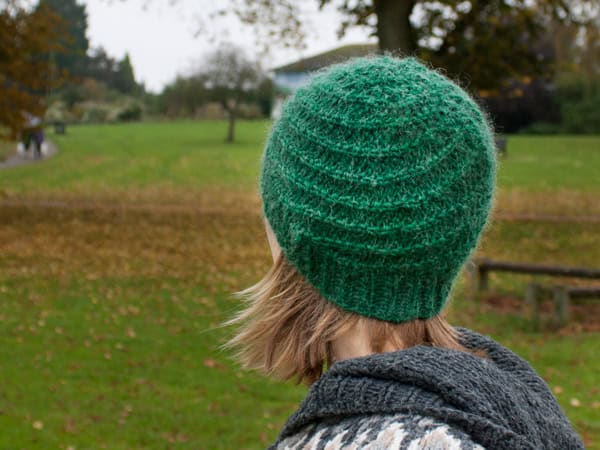 Corkscrew Beanie Chunky Hat Knitting Pattern a person facing away from us wearing a green beanie, knit in stocking stitch with a pattern twisting around in a spiral from the hem to the crown