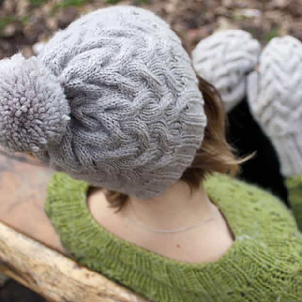 Constantine cable knit hat knitting pattern, shot from above on a person's head. In front out of focus we can see matching mittens.