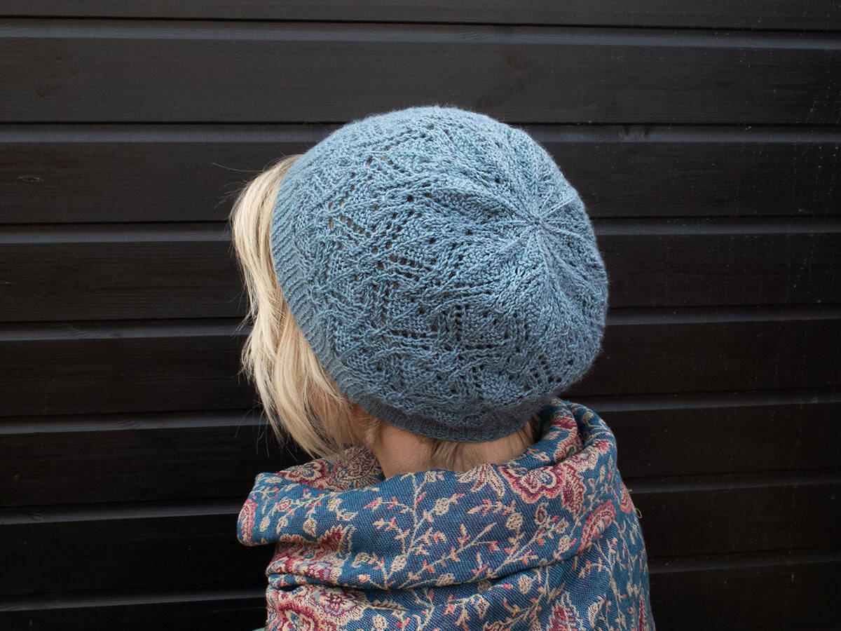 A person with short blonde hair, seen from behind, wears a light blue knitted hat and a patterned shawl in blue, red, and beige tones, standing in front of a dark wooden wall.