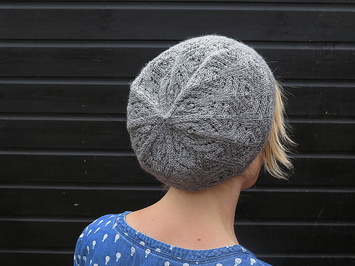A person with blonde hair, seen from behind, wears a knitted grey beret and a blue patterned shirt, standing in front of a black wooden wall.
