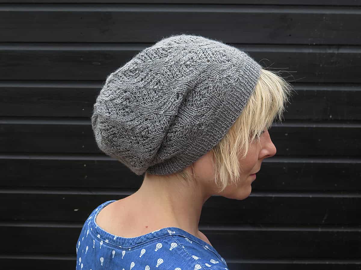 A person with short blonde hair wearing a loose, knitted gray beanie and a blue top with white patterns, standing in front of a black wooden wall. The person is shown in profile, facing right.