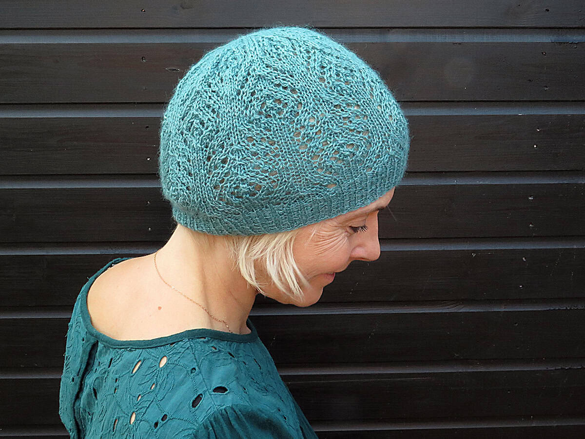 A woman with short blonde hair wears a teal lace knit hat and a matching green top, standing against a dark wooden background and looking down with a gentle smile.