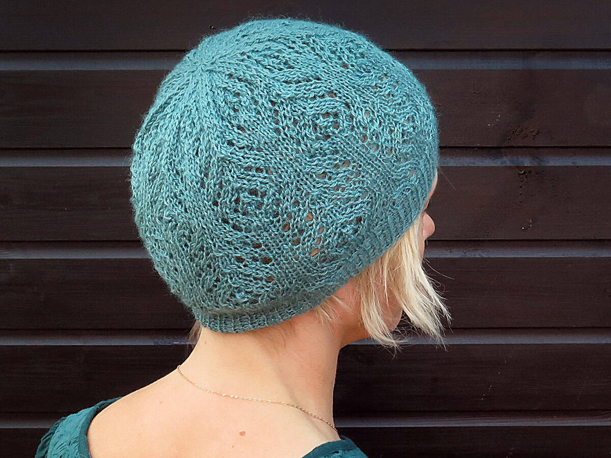 A person with short blonde hair, viewed from behind, wears a lacy teal knit hat and a matching top, standing in front of a dark wooden wall.