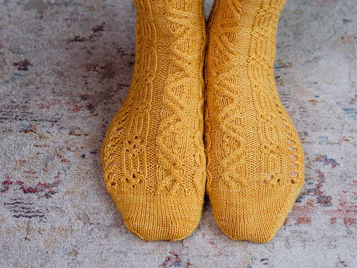 A close-up of two feet wearing bright yellow socks with intricate sock knitting patterns, standing on a light-colored, textured rug with hints of muted colors.