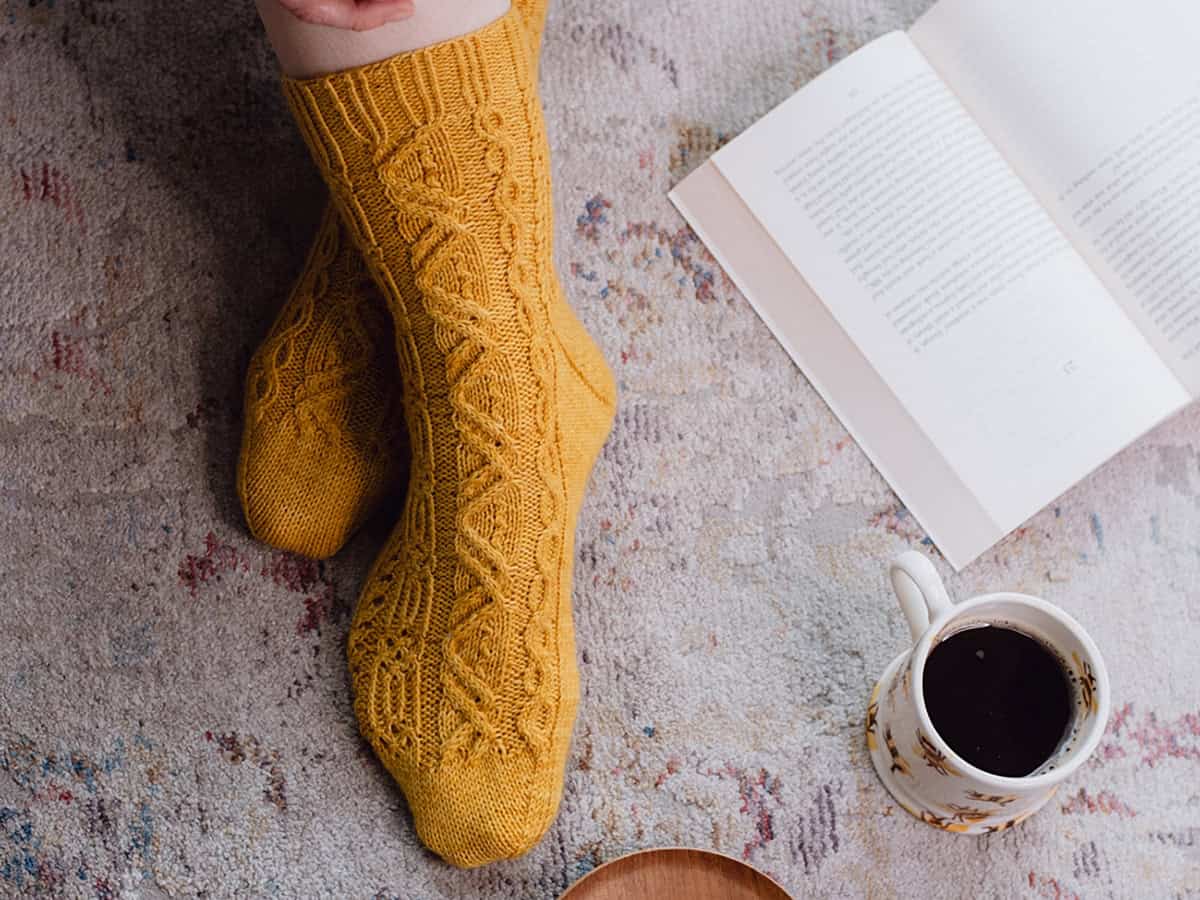Person wearing yellow knitted socks, made with a cozy cable socks pattern, sits on a patterned rug next to an open book and a floral mug filled with coffee.