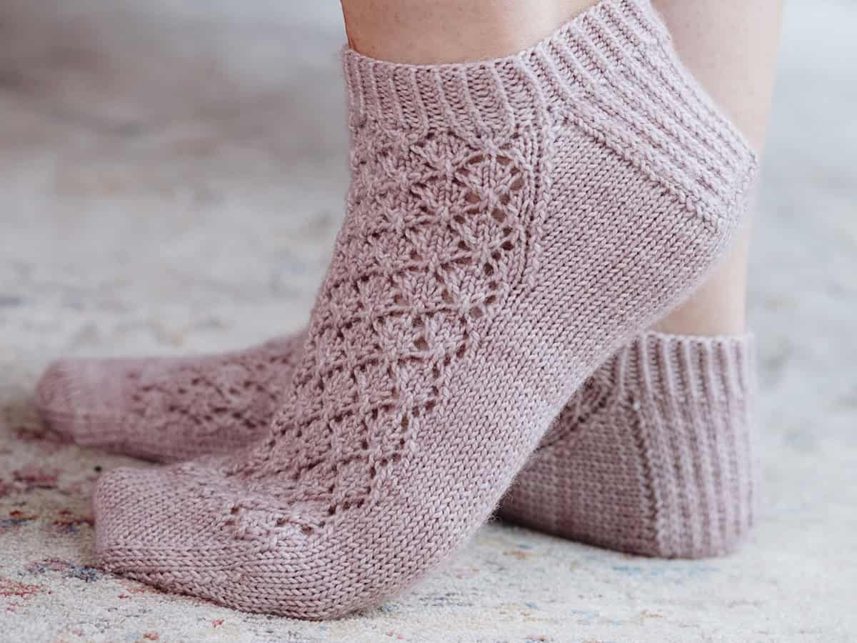 Close-up of two feet wearing light pink, knit shortie socks with a lacy, textured pattern on top and ribbed cuffs, standing on a soft, neutral-colored surface.
