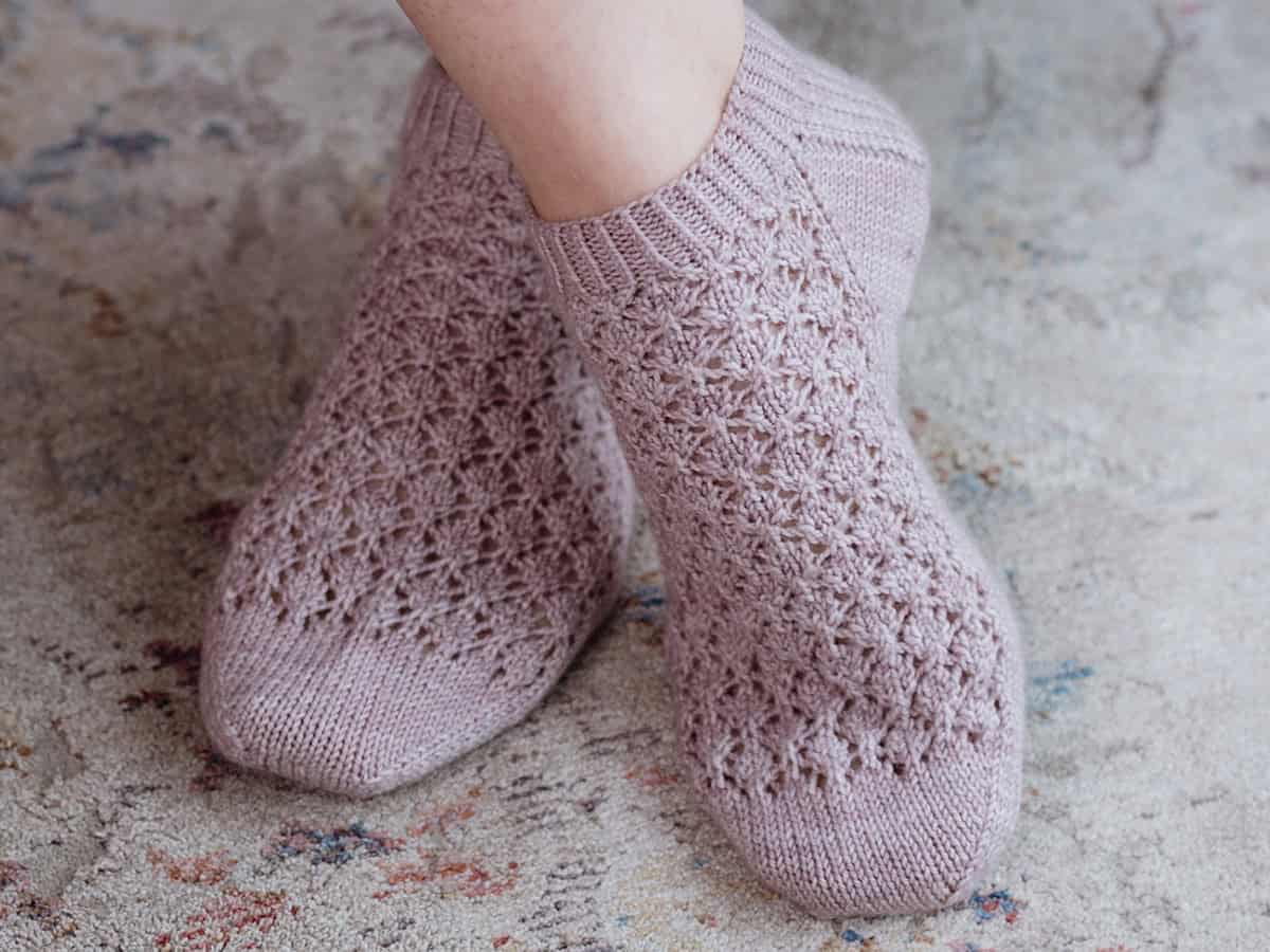Close-up of feet wearing light purple, hand-knitted shortie socks with a delicate lace pattern, standing on a textured, multicolored rug.