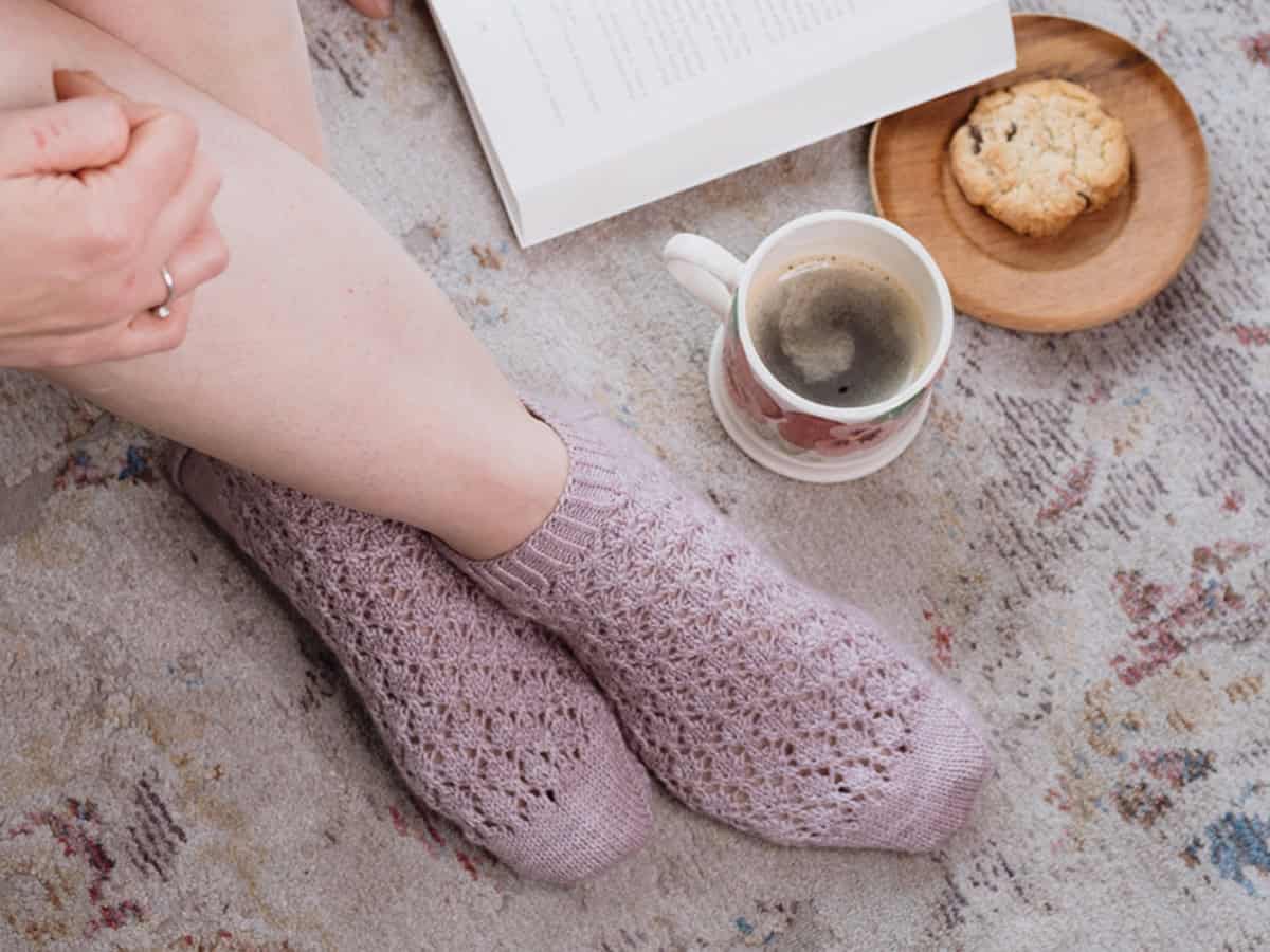 A person wearing cozy shortie socks sits on a patterned rug next to an open book, a cup of coffee, and a cookie on a wooden plate—perfect inspiration for your next knit shortie socks pattern.