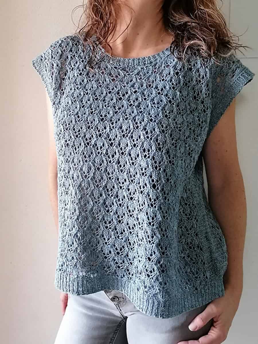 A person wears a light blue, short-sleeved lace tee pattern with a delicate, openwork design, perfect for advanced knitters. Its paired with light-colored jeans against a plain, uncluttered background.