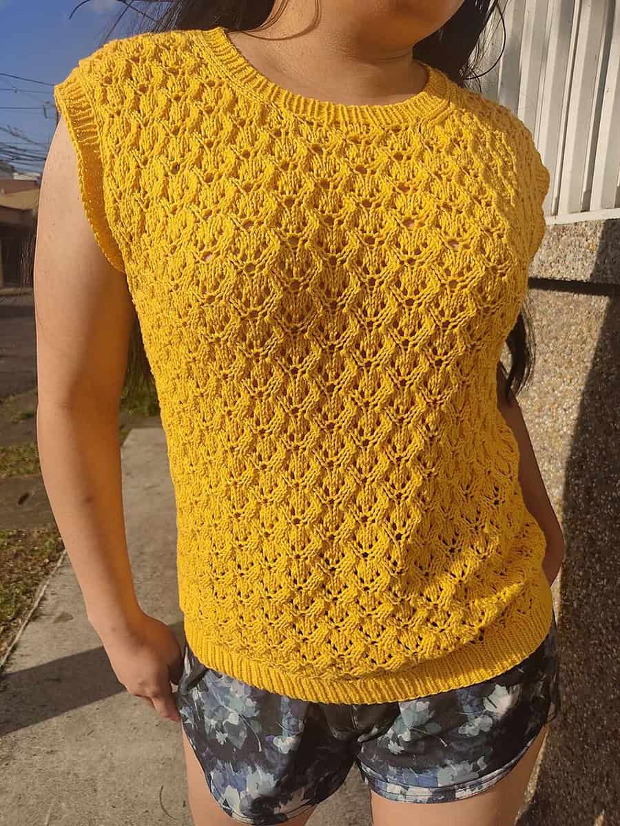 A person wearing a sleeveless, bright yellow knitted top with a textured pattern, paired with dark floral shorts, stands outdoors on a sunny day.