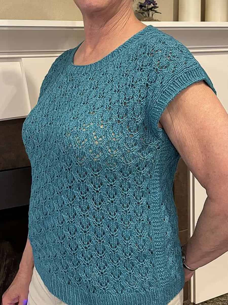 A woman wearing a teal, short-sleeved, lacy knit top stands indoors in front of a white mantel. The top features a delicate openwork pattern.