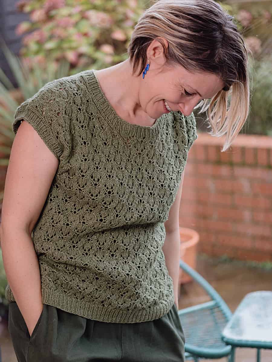A person with short, light brown hair smiles while looking down. They are wearing a green, lacy knit top and dark green pants, standing outdoors near a table and brick wall with plants in the background.