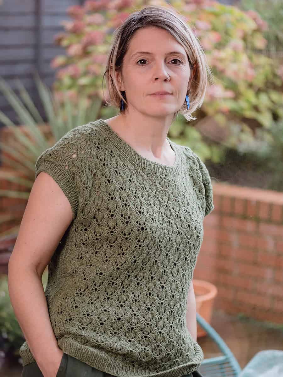 A woman with short, light brown hair stands outdoors, wearing a green knitted short-sleeve top. She looks calmly at the camera, with plants and a brick wall blurred in the background.