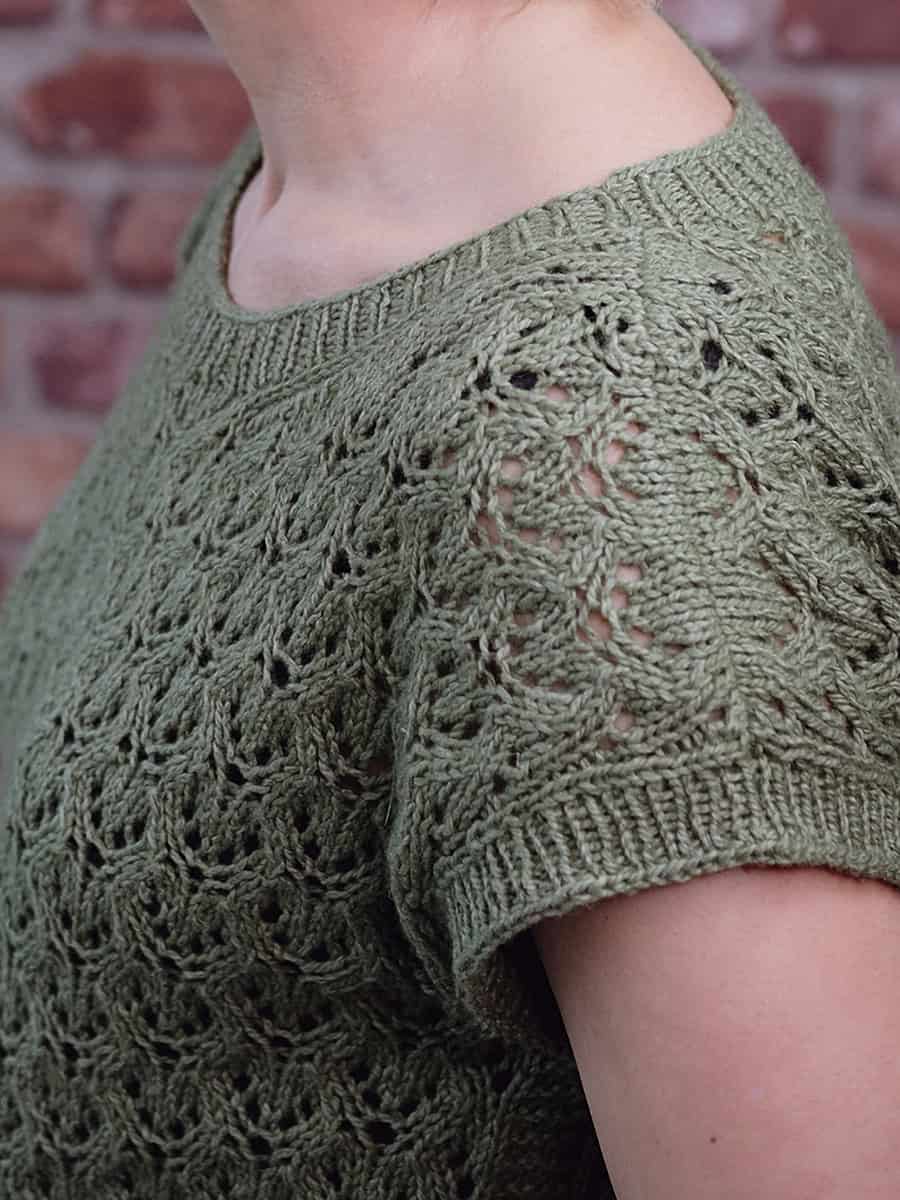 Close-up of a person wearing a green, short-sleeved, knitted top with a lacy, textured pattern. The image is taken from the side, showing the shoulder and upper arm. The background is a blurred brick wall.