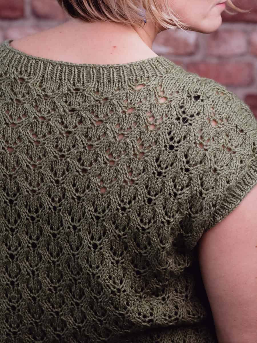 A close-up of a person wearing a green, short-sleeved knit top with an intricate lace pattern on the back, standing in front of a brick wall. The person has short, light brown hair.
