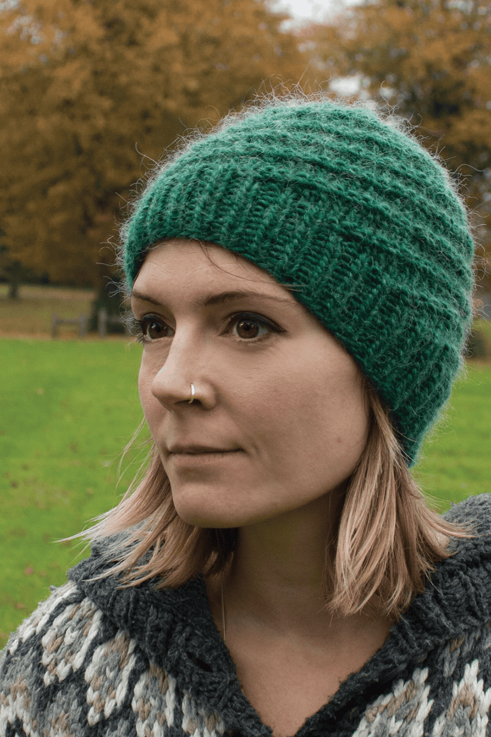 A woman with a nose ring and shoulder-length blonde hair wears a green knitted beanie and a patterned sweater, standing outdoors in a grassy park with autumn trees in the background.