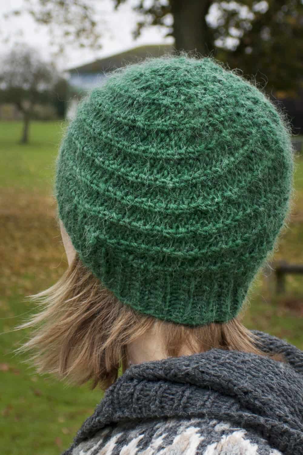 A person with short, light brown hair is wearing a textured, green knitted hat and a grey patterned sweater, standing outdoors in a grassy park with trees in the background. The photo is taken from behind.