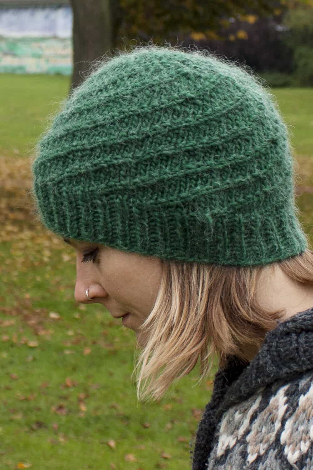 A person with blonde hair wearing a textured green knit hat and a gray sweater is looking downward outdoors, with grass and fallen leaves in the background.