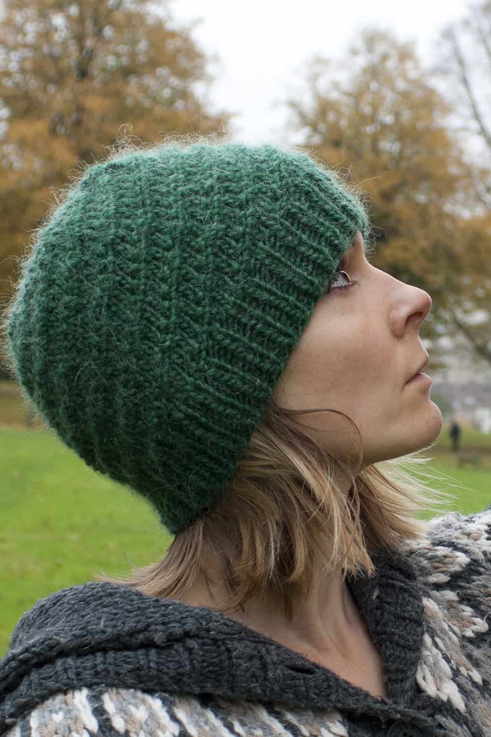 A woman in profile wearing a green knitted beanie and a patterned sweater stands outdoors on a grassy area with autumn trees in the background.
