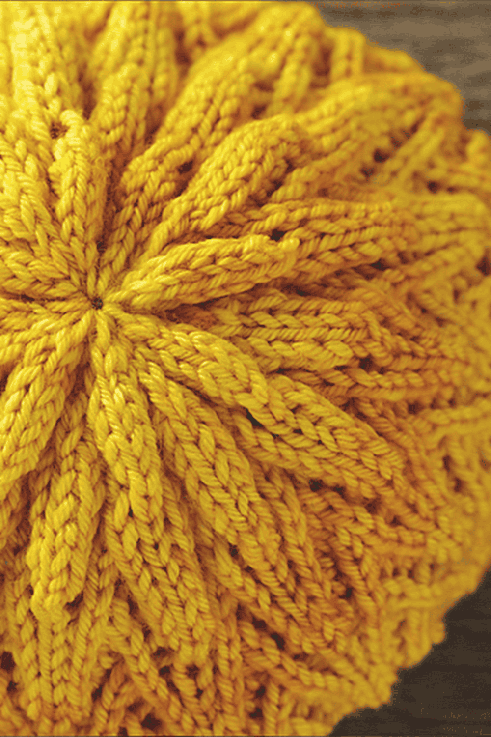 Close-up of a round, hand-knitted yellow hat with a starburst pattern, showcasing textured, radiating ridges and golden yellow tones. This beautiful hat knitting pattern rests on a wooden surface.