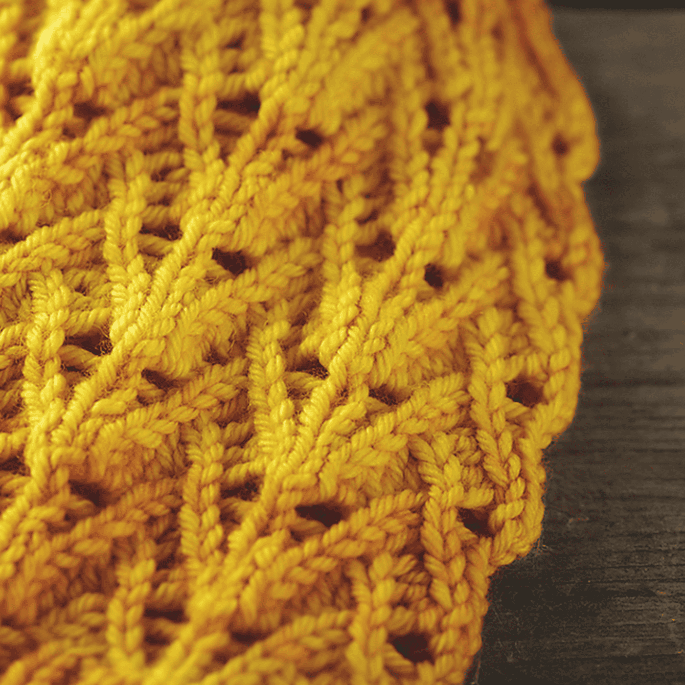 Close-up of a yellow hand-knitted fabric with a textured, lacy hat knitting pattern, lying on a wooden surface. The yarn features subtle variations in shade, creating a warm, vibrant appearance.