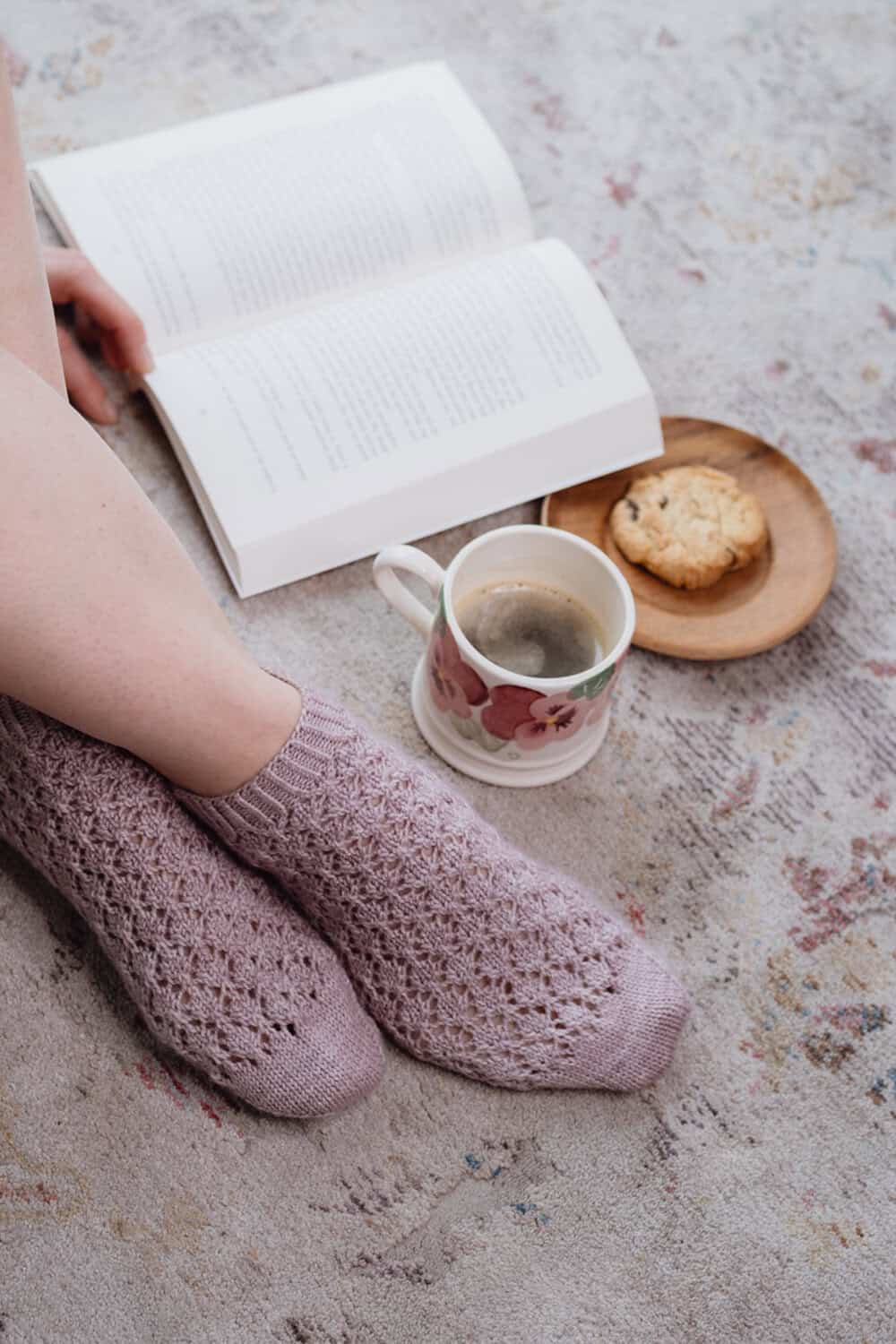 A person wearing light purple knitted socks sits on a carpet with an open book, a cup of coffee, and a cookie on a wooden plate beside them.