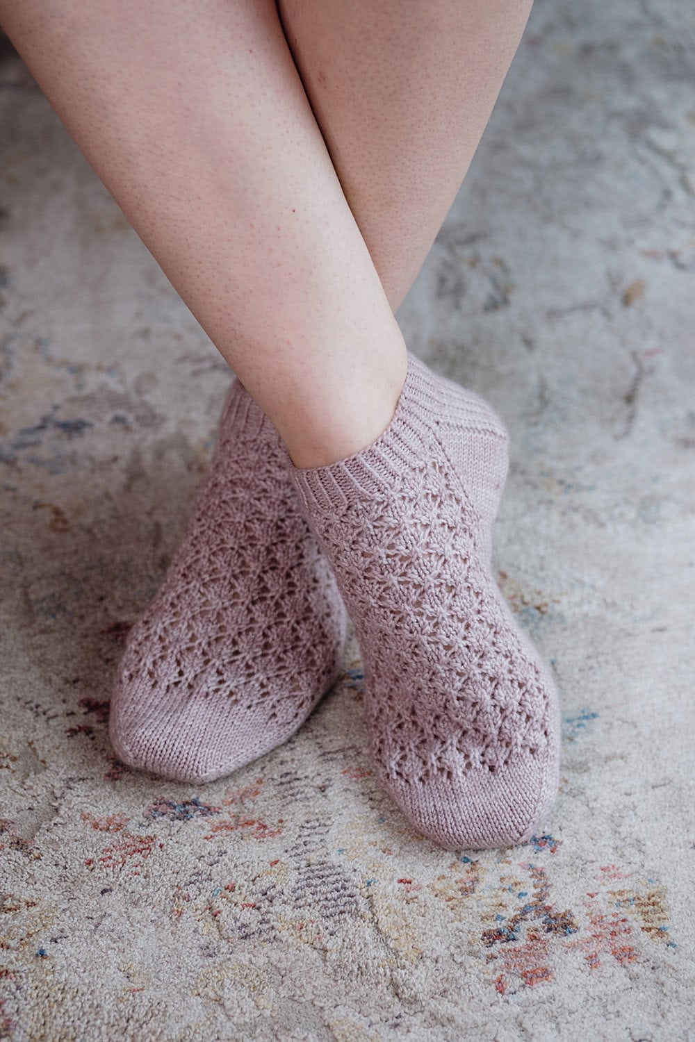 A person with crossed legs is wearing light purple, lacy knit socks while sitting on a soft, patterned rug.