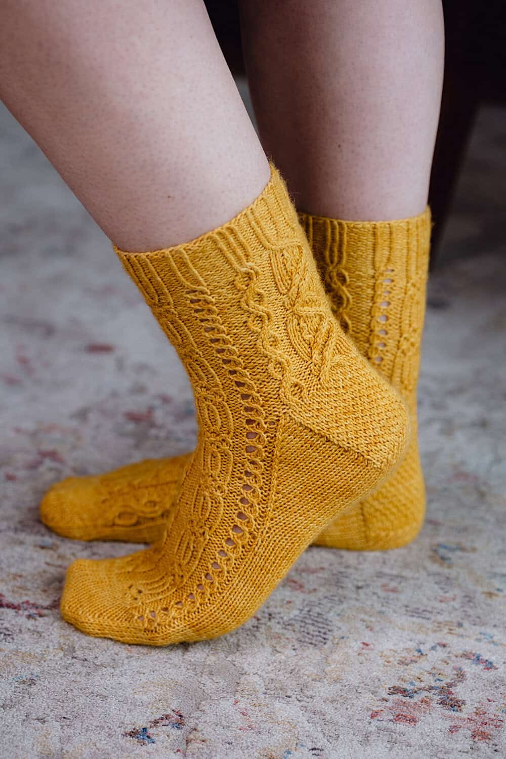 A close-up of a person wearing yellow knitted socks with intricate cable and lace patterns, standing on a light-patterned carpet.