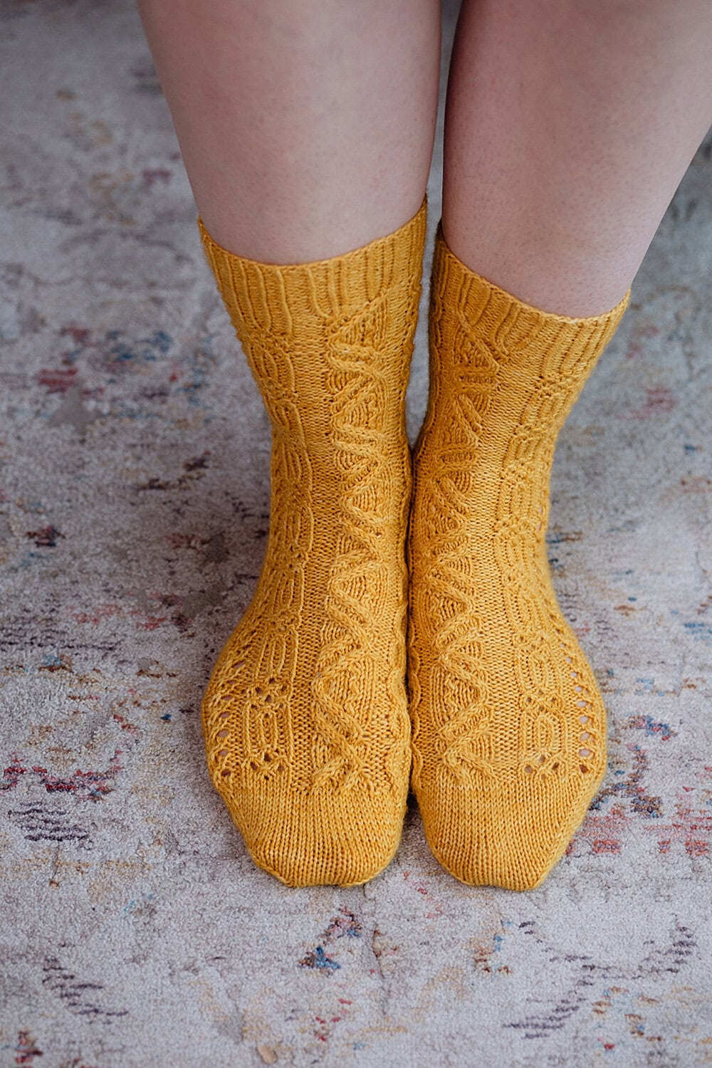 A pair of feet wearing mustard yellow, intricately knitted socks with a cable pattern, standing on a speckled, light-colored carpet.