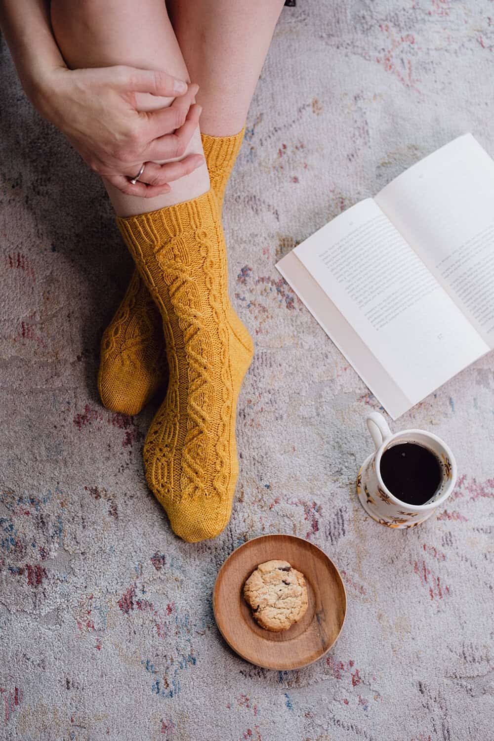 A person in yellow knitted socks sits on a patterned rug with hands resting on their knees, beside an open book, a cup of coffee, and a small plate with a cookie.