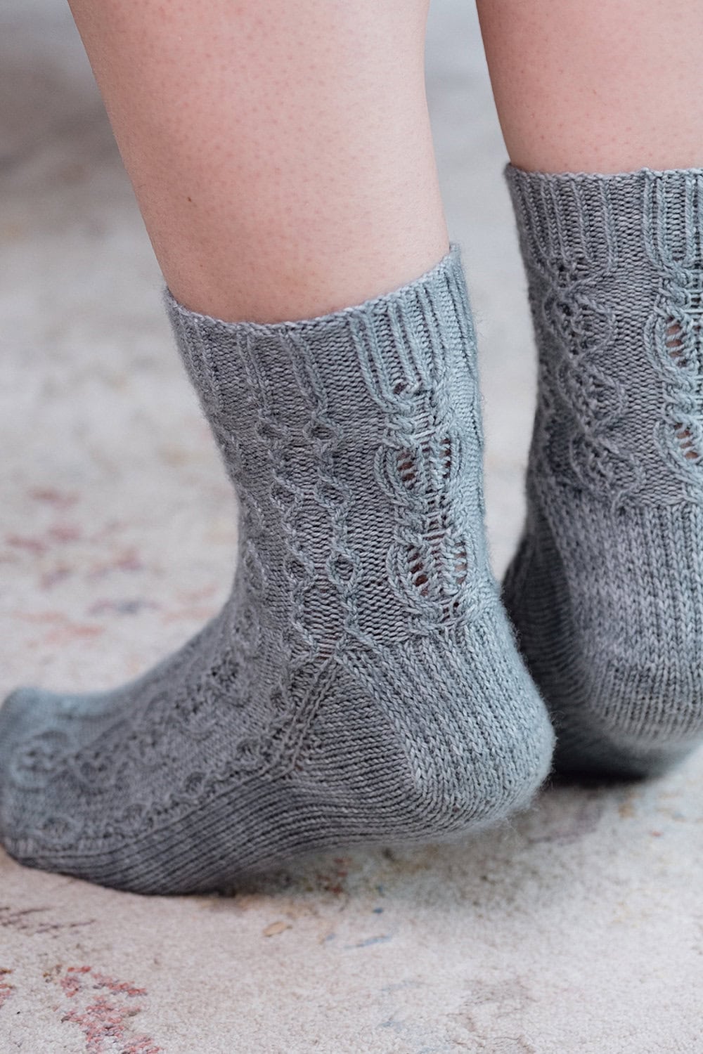 Close-up of a person wearing light gray knitted socks with a textured cable pattern, standing on a soft, speckled surface. Only the lower legs and feet are visible.