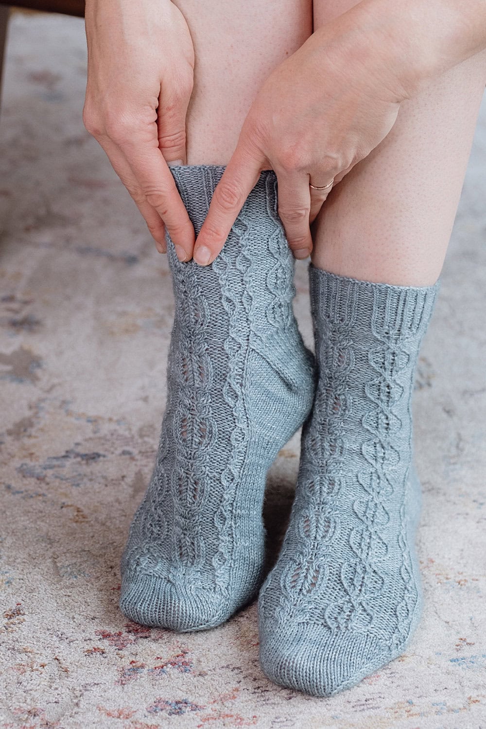 A person wearing textured light blue knitted socks is adjusting one sock while sitting, with hands gently pulling up the fabric over their ankle. The background shows a soft, patterned carpet.