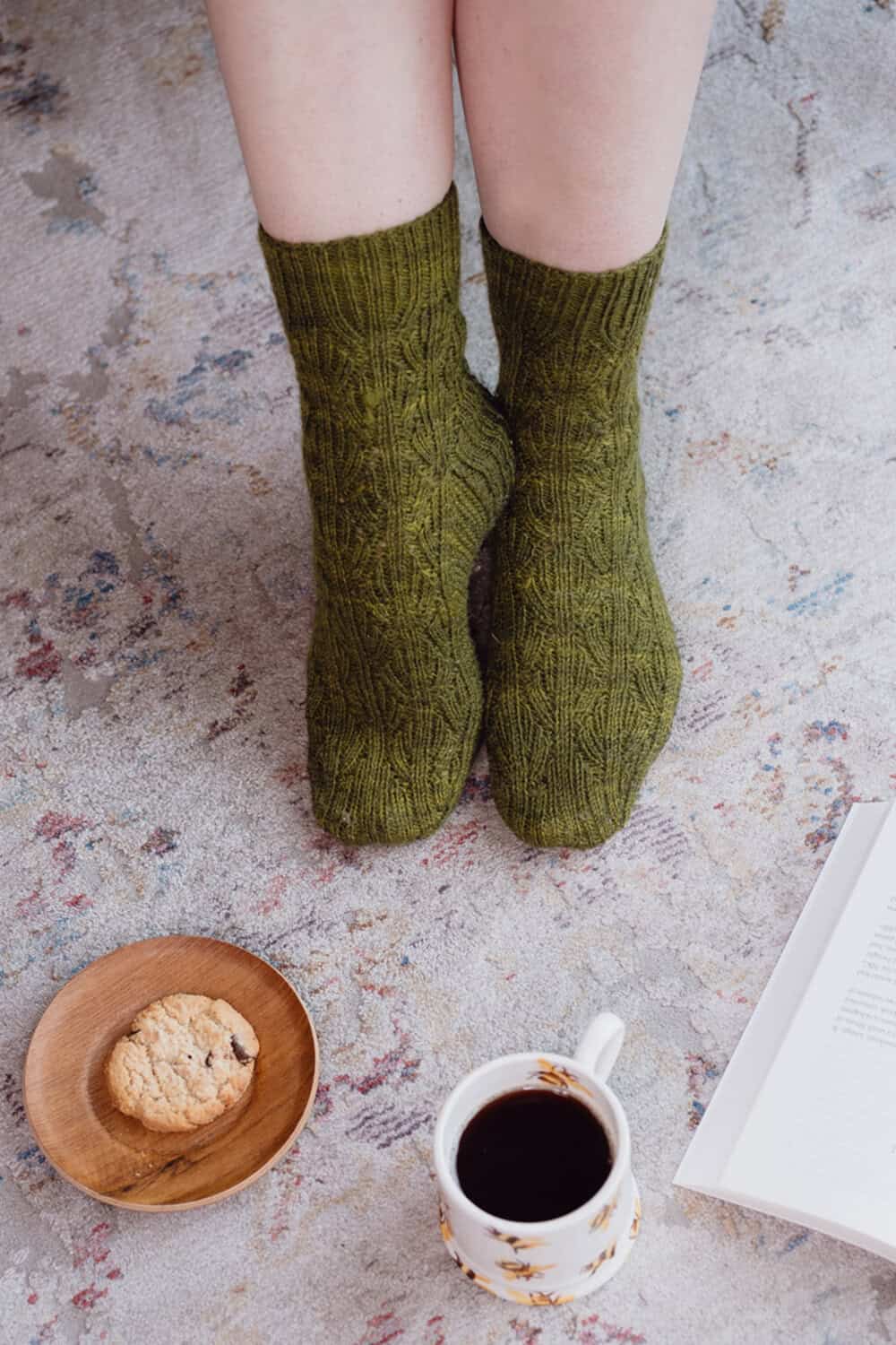 A person wearing green reversible socks sits on a patterned rug with a plate holding a cookie, a cup of coffee, and an open book nearby.