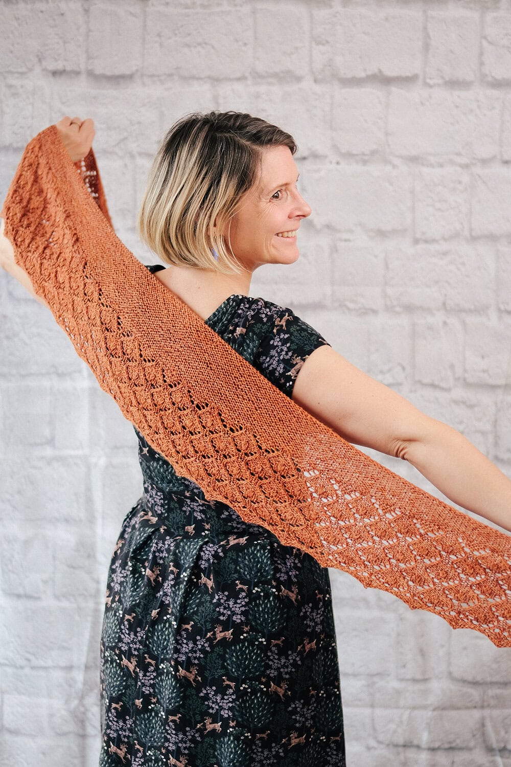 A smiling woman with short blonde hair holds out a rust-colored, lacy knitted shawl behind her back, showing off its pattern. She is wearing a dark, floral dress and standing in front of a light brick wall. The quick knit shawl pattern is Penhale by Faye Perriam-Reed