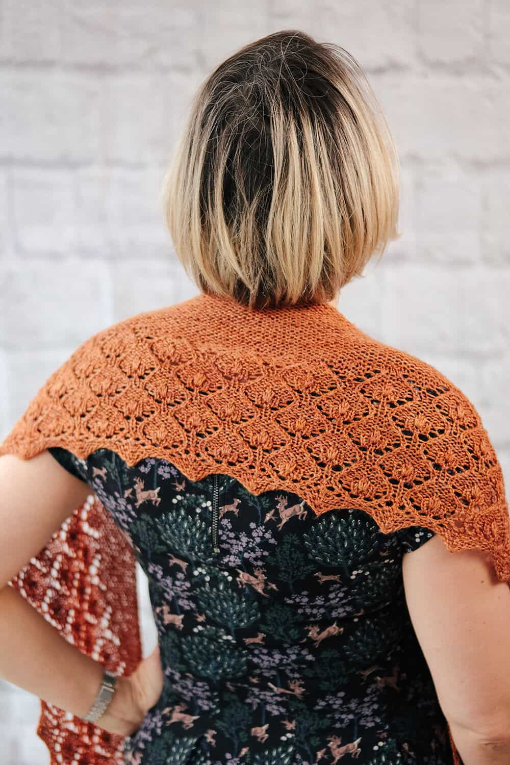 A woman with short blond hair, seen from behind, wears an orange lace knit shawl over a dark floral dress, standing against a light-coloured textured wall. The quick knit shawl pattern is Penhale by Faye Perriam-Reed
