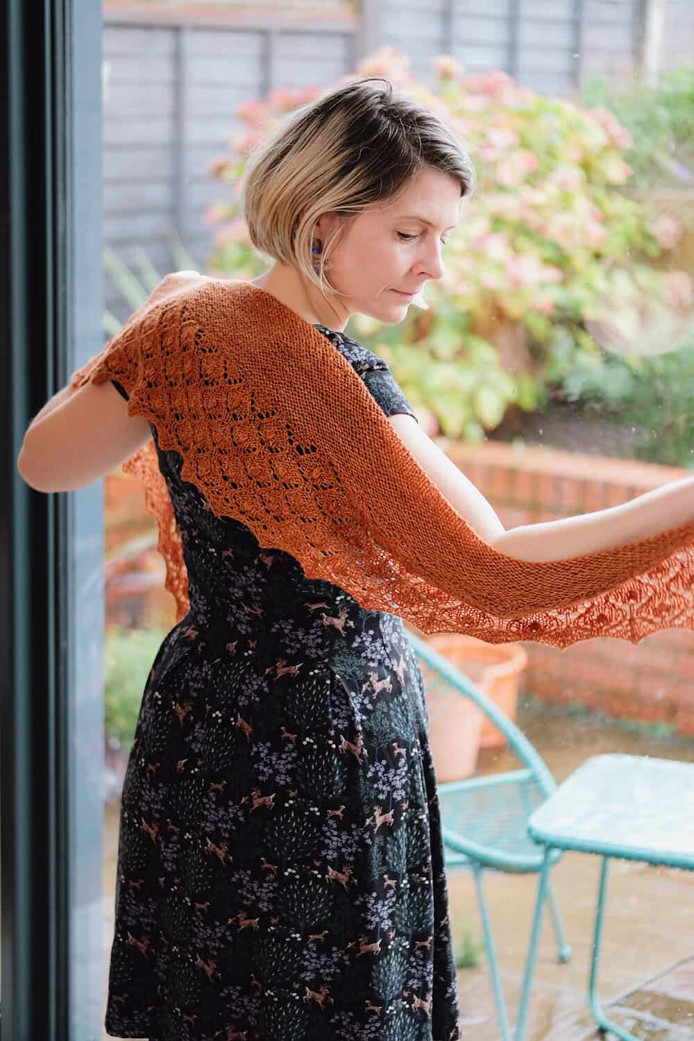 A woman with short blonde hair wearing a black floral dress drapes an orange knitted shawl over her shoulders while standing by a glass door, with a garden and patio furniture visible outside. The quick knit shawl pattern is Penhale by Faye Perriam-Reed