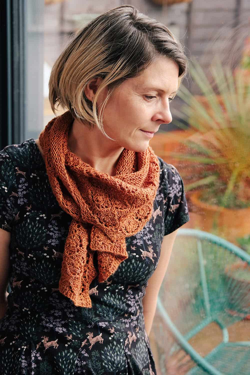 A woman with short blond hair wearing a black dress with an animal print and an orange knitted scarf looks thoughtfully out of a window. A turquoise chair and plants are visible in the background. The quick knit shawl pattern is Penhale by Faye Perriam-Reed and is perfect for summer knitting