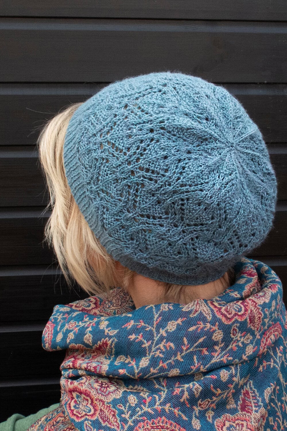 A person with short blonde hair wears a light blue lace hat knitting pattern and a blue floral scarf, standing in front of a dark wooden background.