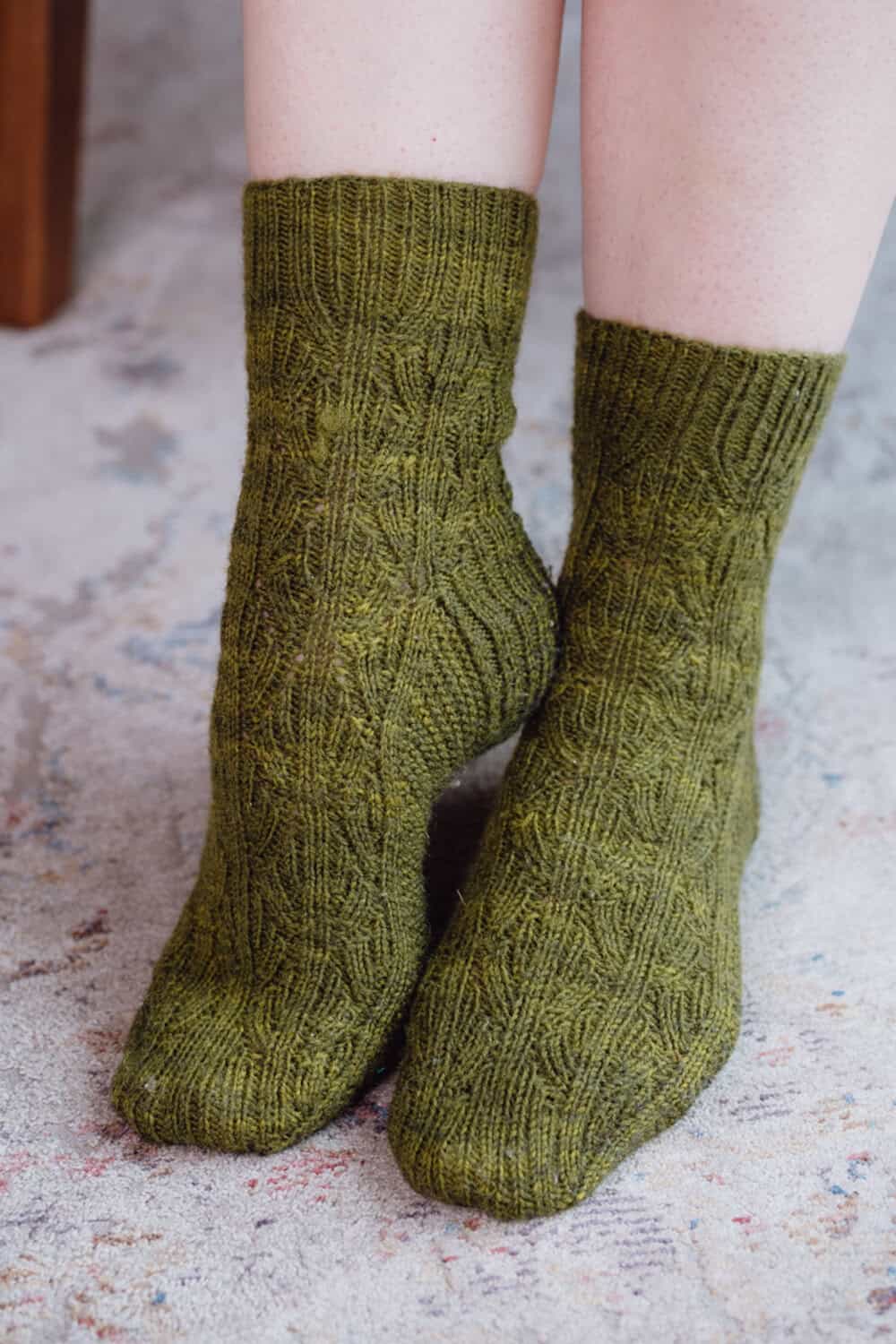 A close-up of two feet wearing green, textured, reversible socks with a hand-knitted cable pattern, standing on a light-colored, patterned carpet.