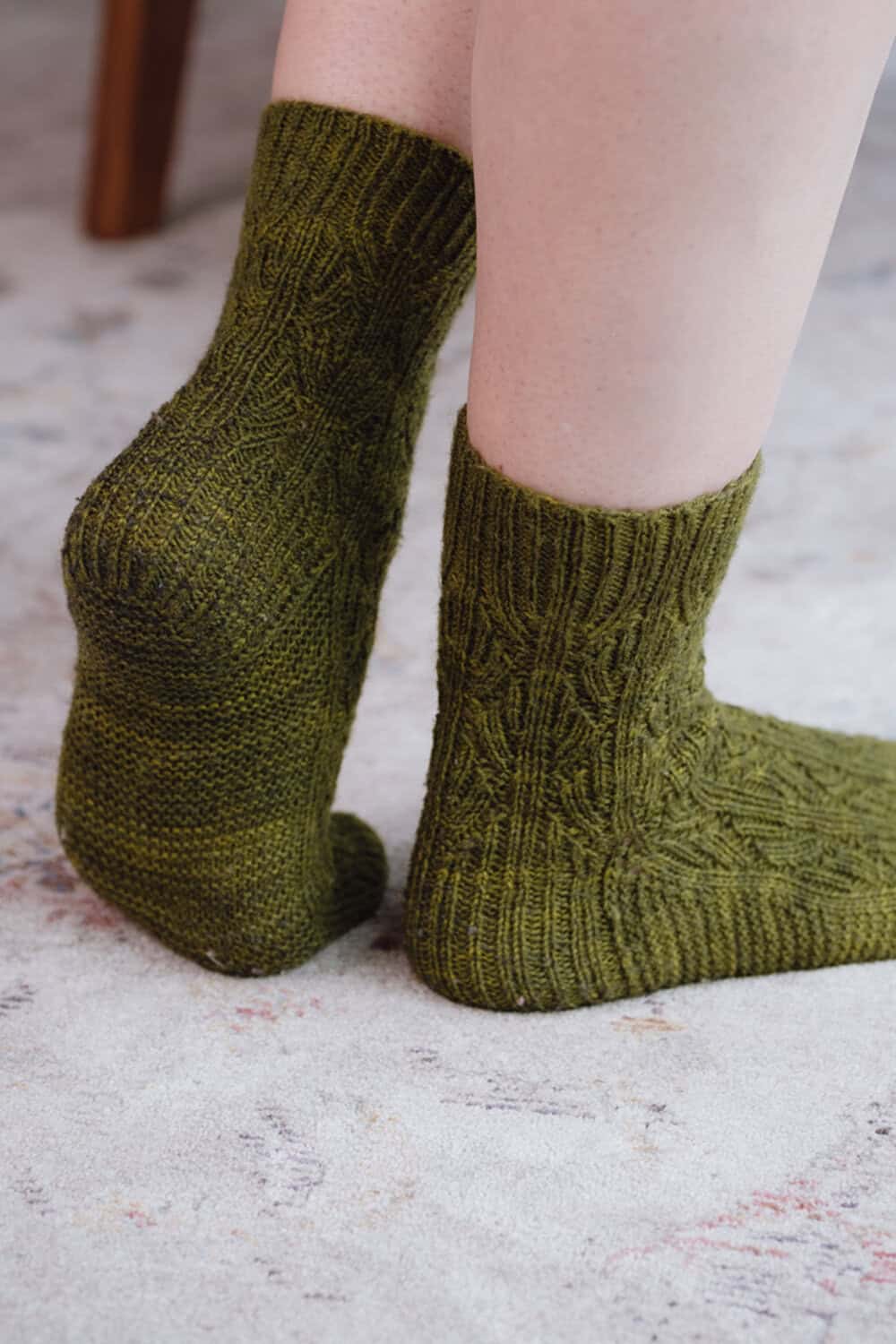 A person wearing olive green reversible socks stands on a light-colored, patterned carpet with their left foot flat and right heel raised.