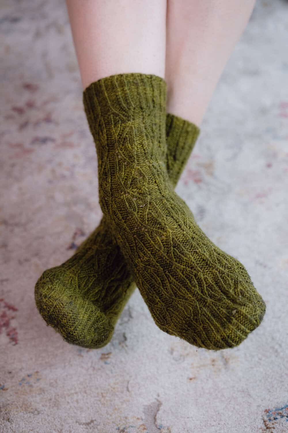 A person with crossed legs wears green, reversible cable knit socks while standing on a patterned, light-colored carpet.