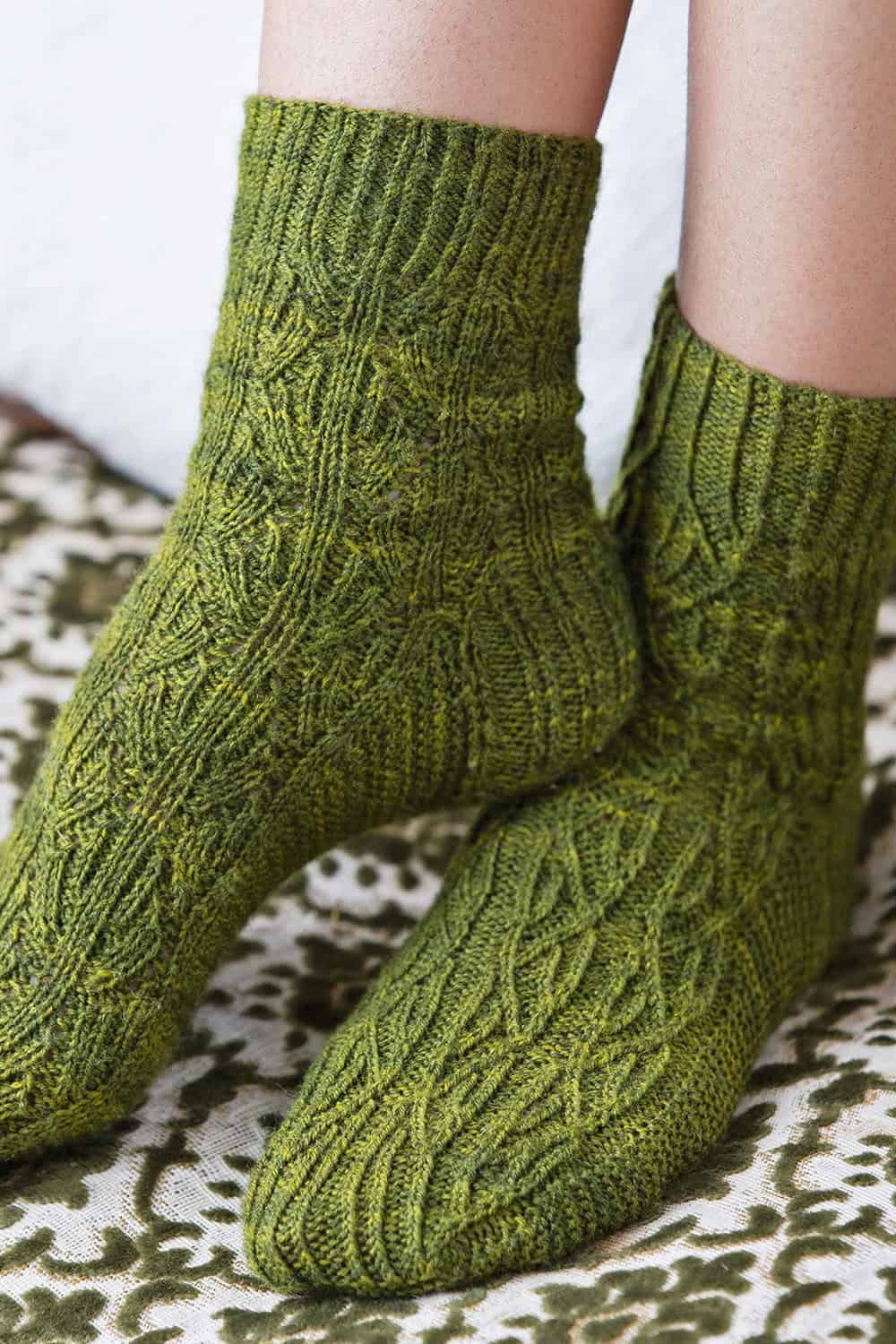 A close-up of two feet wearing hand-knitted, reversible green socks with an intricate cable and leaf pattern. The socks rest on a patterned surface, and part of the persons legs are visible.