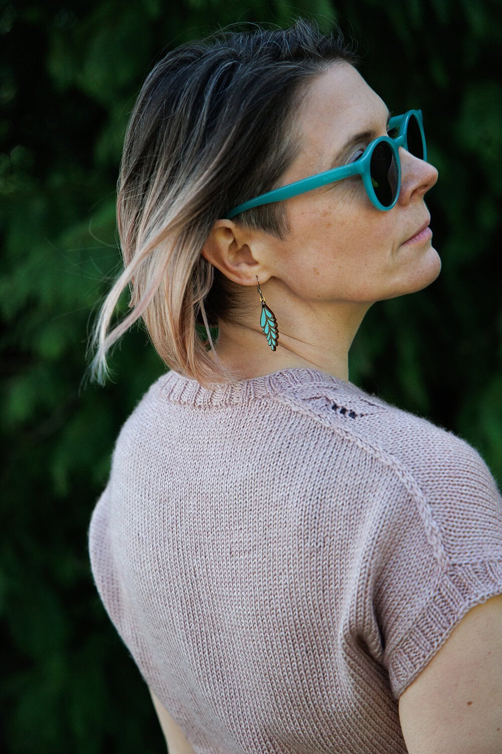 A woman with short, ombre hair wears turquoise sunglasses, a light purple knitted top, and a green feather earring, looking over her shoulder against a blurred green leafy background.