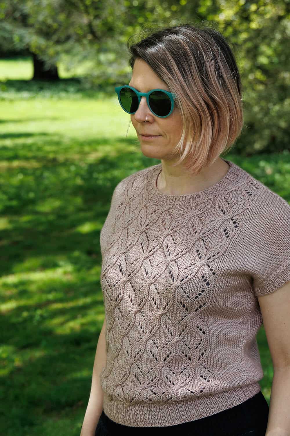 A person wearing green sunglasses and a short-sleeved, pinkish-beige knit top with a lacy diamond pattern stands outside on grass, with trees and sunlight in the background.