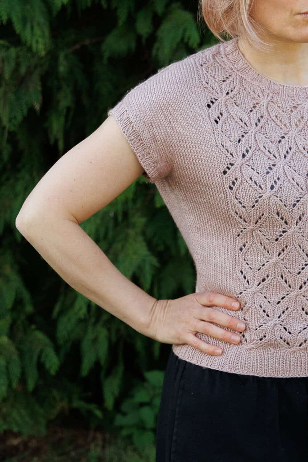 A person with short, light hair stands outdoors, hand on hip, wearing a light pink knit top with intricate lace patterns. Only the upper body is visible, with a green leafy background.