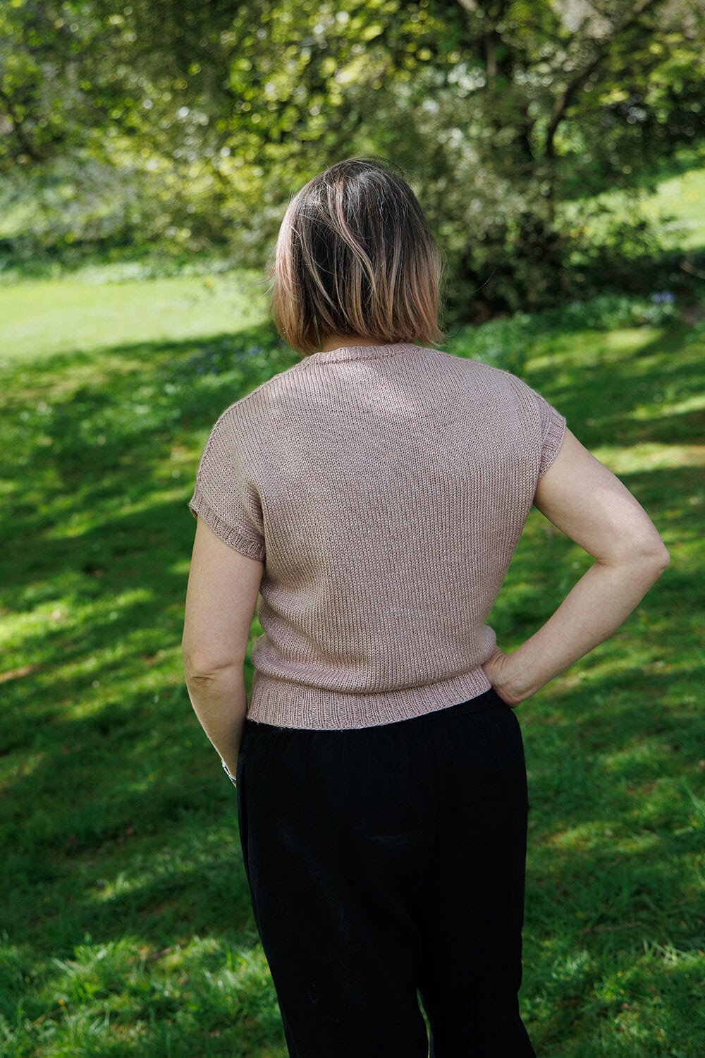 A person with shoulder-length hair stands outdoors on grass, facing away. They wear a short-sleeved, light brown knit top and black pants. Trees and greenery are visible in the background.