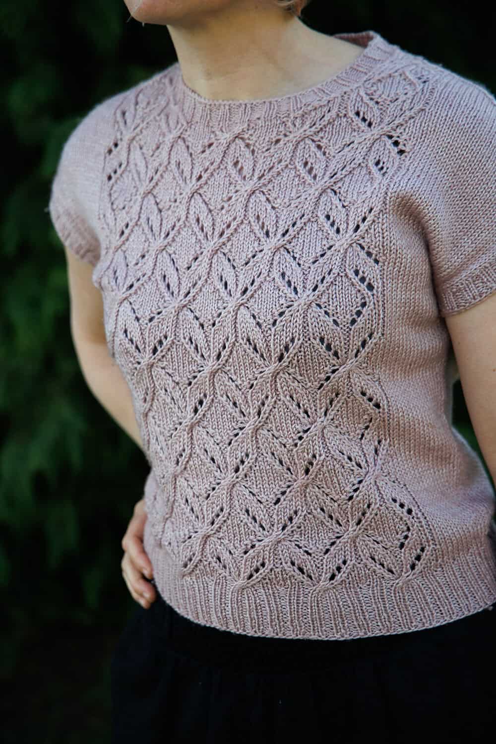 A person wears a short-sleeved, light purple knit sweater with an intricate, lacy leaf pattern on the front, standing outdoors against a dark green, leafy background.