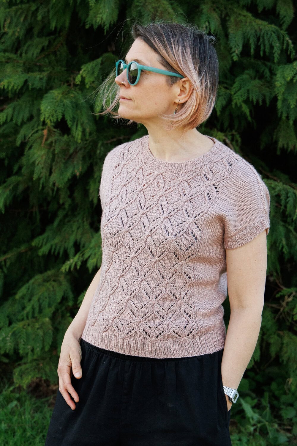 Designer and Tech Editor Faye Perriam-Reed is wearing sunglasses and modelling her pale pink Tywardreath lace summer knitted top. She stands outdoors in front of green foliage, looking to the left.