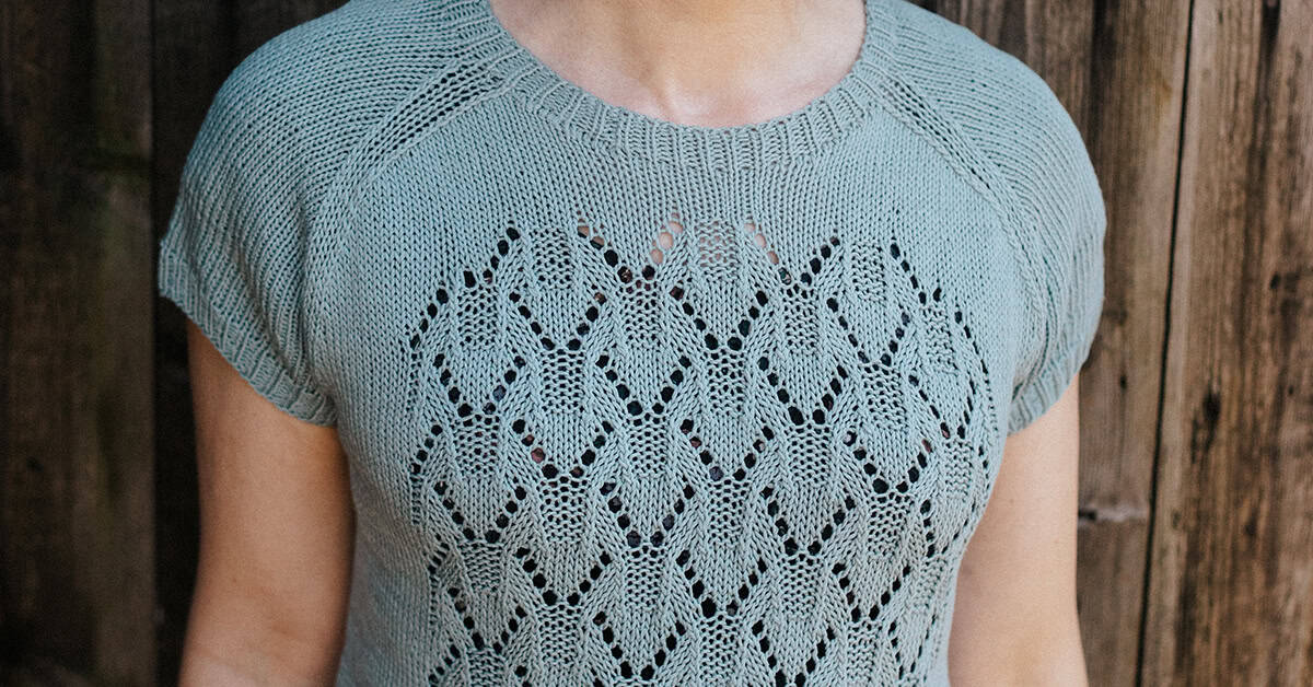 A person wearing a light green short-sleeve knit top with a diamond-shaped lace pattern on the front stands in front of a wooden background.