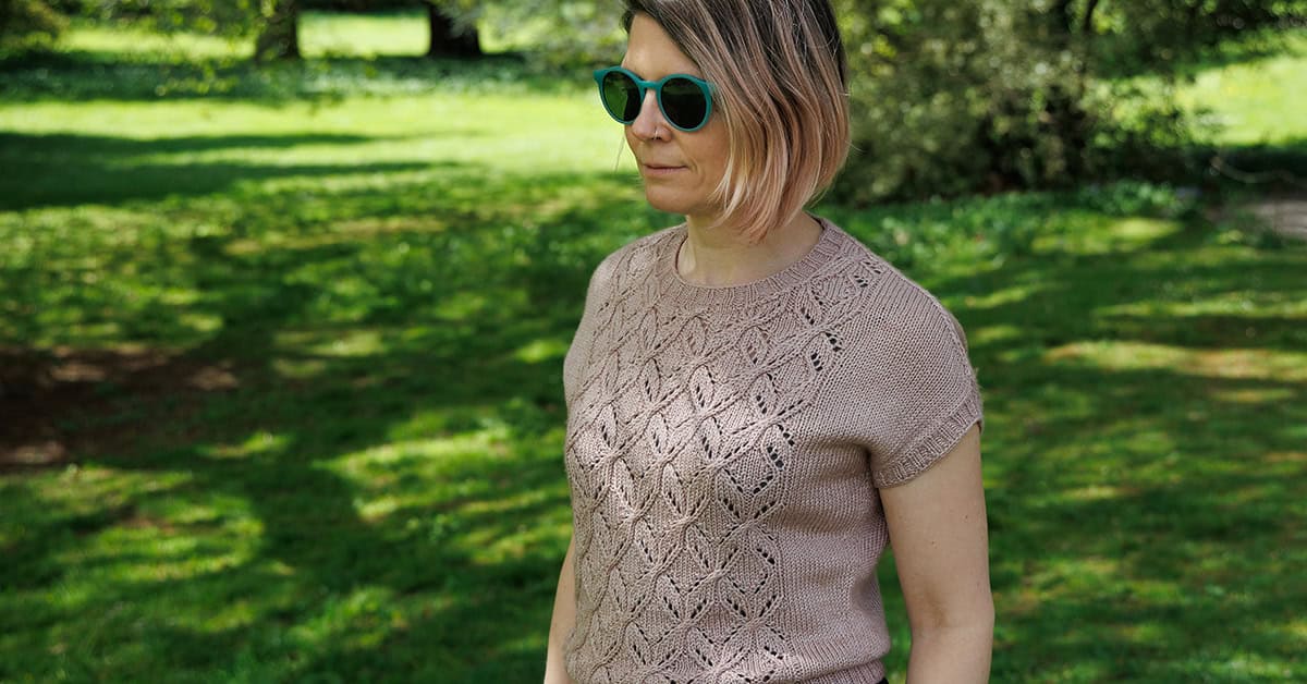 A person wearing sunglasses and a short-sleeved, light brown knitted sweater with a diamond lace pattern stands outside on a sunny day, with green grass and trees in the background. Design for the top is Tywardreath Tee summer knitting pattern by Faye Perriam-Reed